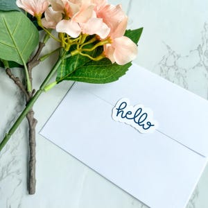 May include: A white envelope with a blue "hello" sticker on it. A bouquet of pink flowers with green leaves is in the background.