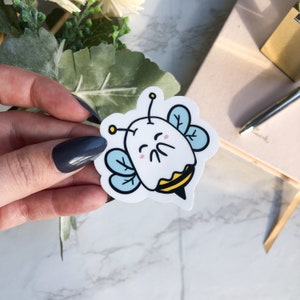 May include: A white bee sticker with blue wings and yellow stripes. The bee has a cute, cartoon-like face with closed eyes and a small smile.