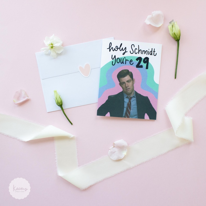 May include: A greeting card featuring a printed illustration of a man in a suit and tie against a wavy, multi-colored background. The text reads, "holy Schmidt You're 29." The card is accompanied by a white envelope with a small heart sticker.