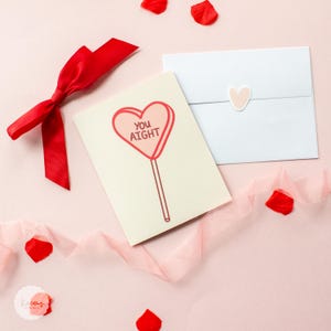 May include: A Valentine's Day card with a pink heart-shaped lollipop with the text "You Aight" on it. The card is on a pink background with a red ribbon and pink petals.