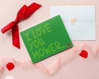 May include: A green card with the text "I love you mower" in a playful font. The card is surrounded by pink tissue paper, red rose petals, and a white envelope with a pink heart sticker.