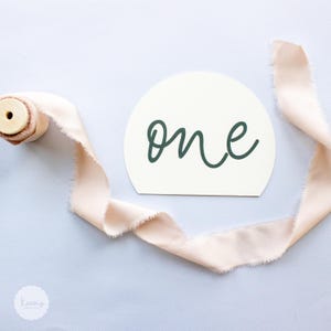 May include: A white, semi-circular card with the word "one" written in a cursive font in dark green. The card is lying on a light pink ribbon.