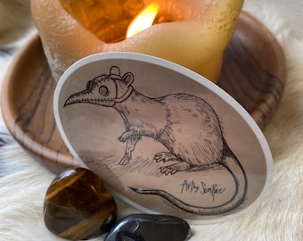 May include: An oval sticker featuring a detailed pen and ink illustration of a rat wearing a plague doctor mask. The sticker is placed on a white surface with two polished stones and a lit candle in the background. The text "Art by SunRae" is visible.