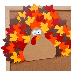 Puede incluir: Un recorte artesanal de pavo hecho de cartulina color canela con una car&uacute;ncula roja, un pico naranja y dos ojos blancos con pupilas negras. El pavo presenta una gran cola en abanico hecha de hojas de arce de papel superpuestas en tonos rojo, naranja, amarillo y marr&oacute;n.