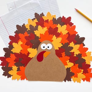 Puede incluir: Un pavo de papel con cuerpo marr&oacute;n, ojos blancos, pico naranja y car&uacute;ncula roja. El pavo tiene una gran cola en abanico hecha de numerosas hojas de papel superpuestas en tonos amarillo, naranja, rojo y marr&oacute;n, dispuestas en un patr&oacute;n circular.