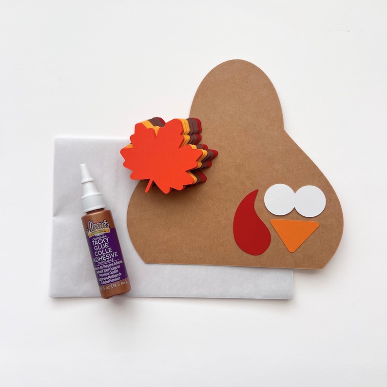 Puede incluir: Un kit de manualidades con una silueta de pavo en cartulina marr&oacute;n, una pila de hojas de arce de papel en naranja y rojo, formas de papel blanco y naranja para la cara y un frasco de pegamento Aleene's sobre un sobre blanco.