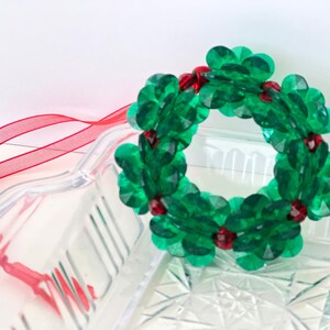 Pode incluir: Um enfeite de Natal de grinalda de contas verdes com contas vermelhas. A grinalda est&aacute; pendurada em uma fita vermelha.