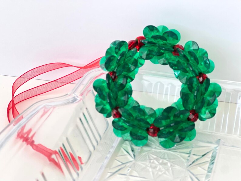 Pode incluir: Um enfeite de Natal de grinalda de contas verdes com contas vermelhas. A grinalda est&aacute; pendurada em uma fita vermelha.