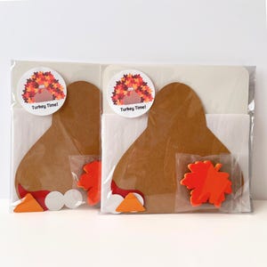 Puede incluir: Dos kits de manualidades en envases de pl&aacute;stico transparente, cada uno con un recorte de cartulina marr&oacute;n en forma de pavo, una pegatina circular con una ilustraci&oacute;n de pavo y el texto Turkey Time, y una pieza de espuma naranja con forma de hoja de arce.
