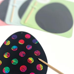 May include: Black scratch art egg with rainbow-colored polka dots and a wooden stylus. The egg-shaped scratch art is on a white surface, with other scratch art sheets in pink, blue, and green in the background.