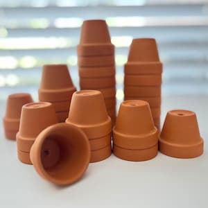 May include: A collection of small, terracotta-colored clay pots. The pots are stacked in varying heights, with some arranged individually. The unglazed pots have a classic, tapered design, perfect for planting small herbs or succulents.