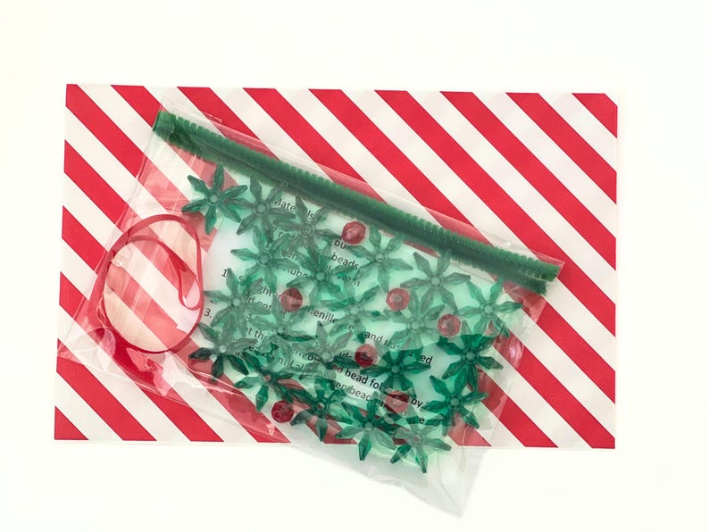 Pode incluir: Um saco de pl&aacute;stico transparente com contas verdes em forma de estrela, um limpador de cachimbo vermelho e um cart&atilde;o branco com o texto "Celebrate with you!" O saco est&aacute; sobre uma superf&iacute;cie listrada vermelha e branca.