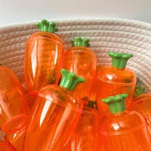 May include: A collection of orange, carrot-shaped containers with green tops, nestled in a woven basket. The transparent plastic allows a glimpse of the contents. The containers are likely for Easter or spring-themed treats.