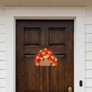 Puede incluir: Una decoraci&oacute;n para puerta con forma de pavo que presenta un cuerpo color canela, una car&uacute;ncula roja y una gran cola en forma de abanico hecha de coloridas hojas de oto&ntilde;o en tonos naranja, amarillo y rojo. El pavo tiene dos ojos blancos.