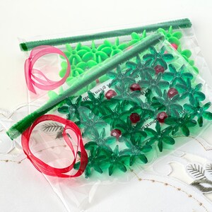 Pode incluir: Um saco de pl&aacute;stico transparente cheio de decora&ccedil;&otilde;es de &aacute;rvore de Natal de pl&aacute;stico verde e vermelho. As decora&ccedil;&otilde;es t&ecirc;m a forma de uma estrela e uma folha de azevinho. O saco &eacute; amarrado com uma fita vermelha.