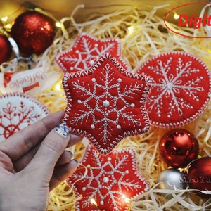May include: Red felt star ornaments with white embroidered snowflakes. The ornaments are in a box with shredded paper and string lights. The text "Digital" is written in red on a white background.