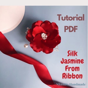 Puede incluir: Una flor de seda roja con una perla y un adorno plateado. La flor está sobre un fondo azul con una cinta roja. El texto "Tutorial PDF Silk Jasmine From Ribbon" está en el lado derecho de la imagen. El texto "NhanDo Handmade" está en la parte inferior de la imagen.