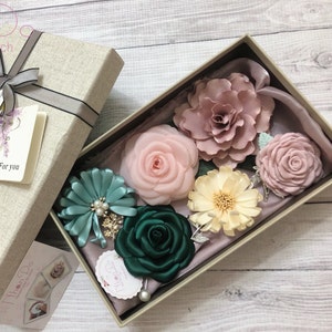 May include: A set of six fabric flower hair clips in a gift box. The flowers are in shades of pink, green, and cream. The box is lined with a light pink fabric.