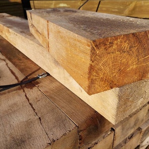 May include: Close-up of a stack of rough-hewn, light brown wooden beams. The beams are stacked on top of each other, and the grain of the wood is visible. The beams are likely used for construction or landscaping.