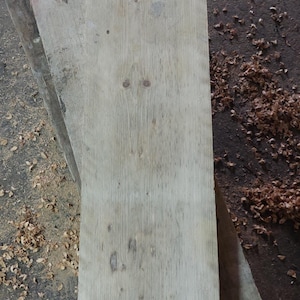 May include: A weathered, light grey wooden plank with a rough texture. The plank is lying on a surface covered in sawdust and wood chips.