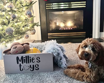 May include: A brown and white dog sits in front of a fireplace with a Christmas tree in the background. A wooden toy box with the text "Millie's toys" is on the floor in front of the dog.