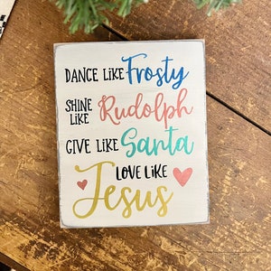May include: White wooden sign with the text "Dance like Frosty, Shine like Rudolph, Give like Santa, Love like Jesus" in colorful lettering. The sign has two pink hearts on either side of the word "Jesus".