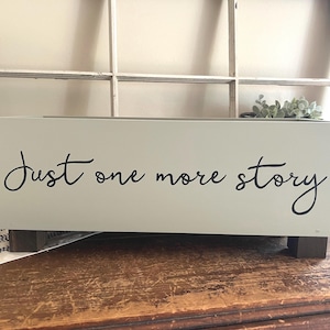 May include: A rectangular, light green wooden box with the words "Just one more story" in black script. The box sits on two dark brown wooden feet. A small plant is visible in the background.