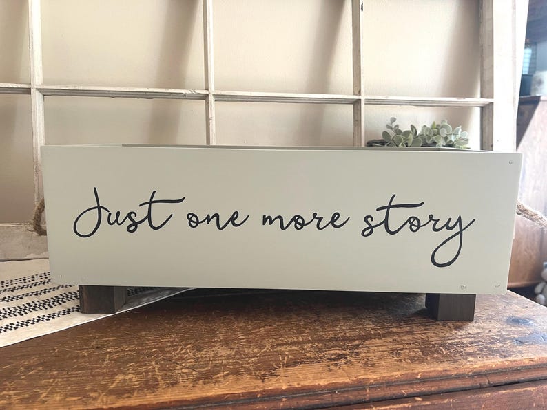 May include: A rectangular, light green wooden box with the words "Just one more story" in black script. The box sits on two dark brown wooden feet. A small plant is visible in the background.