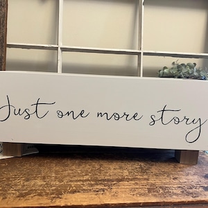 May include: A white rectangular wooden box with the text "Just one more story" in black script. The box has small wooden feet and rope handles. The box is on a wooden surface.