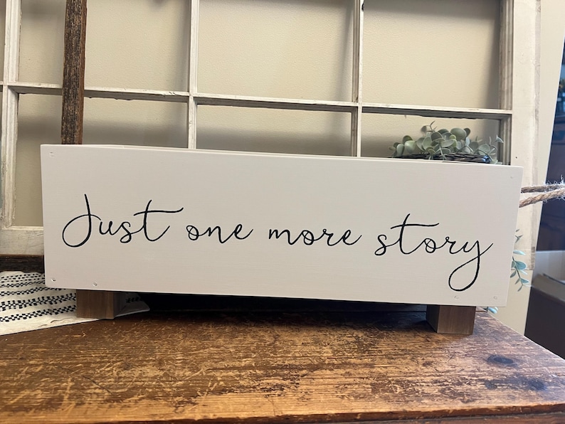 May include: A white rectangular wooden box with the text "Just one more story" in black script. The box has small wooden feet and rope handles. The box is on a wooden surface.