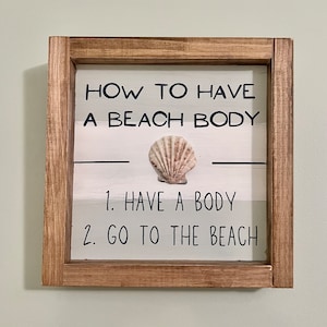 May include: Wooden framed sign with the text "HOW TO HAVE A BEACH BODY" and the steps "1. HAVE A BODY 2. GO TO THE BEACH". A seashell is centered on the sign. The frame is brown, and the background is white and light gray.