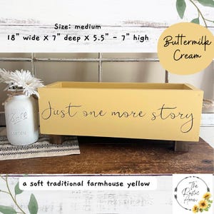May include: A medium-sized, soft yellow wooden box with the text "Just one more story" in black script. The box is 18 inches wide, 7 inches deep, and 5.5 to 7 inches high. A mason jar with white flowers sits to the left.