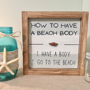May include: A wooden framed sign with the text "HOW TO HAVE A BEACH BODY" and the steps "1. HAVE A BODY 2. GO TO THE BEACH". A blue glass jar with a starfish and a small jar with sand and shells are also visible.