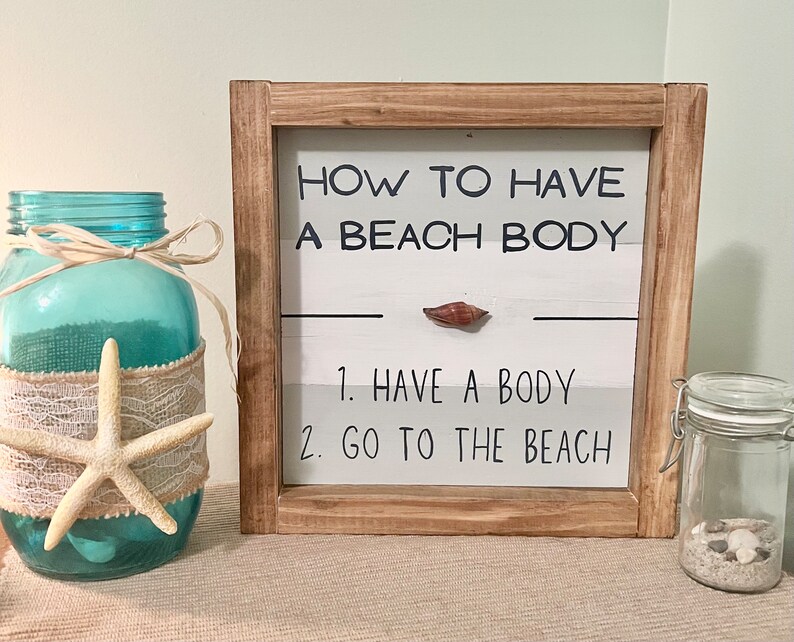 May include: A wooden framed sign with the text "HOW TO HAVE A BEACH BODY" and the steps "1. HAVE A BODY 2. GO TO THE BEACH". A blue glass jar with a starfish and a small jar with sand and shells are also visible.