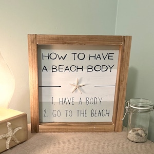 May include: A wooden framed sign with the text "HOW TO HAVE A BEACH BODY" and the steps "1. HAVE A BODY 2. GO TO THE BEACH". A starfish is affixed to the sign. A glass jar with sand and shells is to the right.