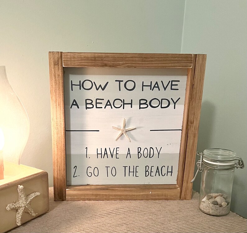 May include: A wooden framed sign with the text "HOW TO HAVE A BEACH BODY" and the steps "1. HAVE A BODY 2. GO TO THE BEACH". A starfish is affixed to the sign. A glass jar with sand and shells is to the right.
