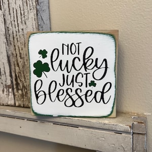May include: Square wooden sign with the words "NOT lucky JUST blessed" in black script. The sign has a white background with green trim and shamrock accents. A decorative item for St. Patrick's Day.