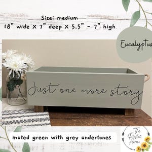 May include: A muted green wooden box with grey undertones, measuring 18 inches wide, 7 inches deep, and 5.5 to 7 inches high. The box has the text "Just one more story" in black script. Features rope handles and wooden feet.