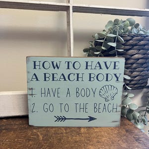 May include: A distressed, rectangular wooden sign with the text "HOW TO HAVE A BEACH BODY" in navy blue. The sign lists two steps: "1. HAVE A BODY" and "2. GO TO THE BEACH". A seashell graphic and an arrow are also included.