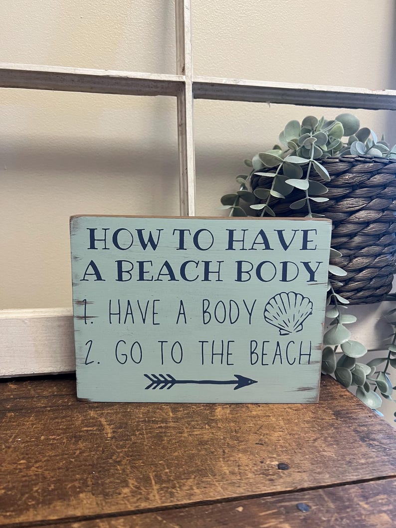 May include: A distressed, rectangular wooden sign with the text "HOW TO HAVE A BEACH BODY" in navy blue. The sign lists two steps: "1. HAVE A BODY" and "2. GO TO THE BEACH". A seashell graphic and an arrow are also included.