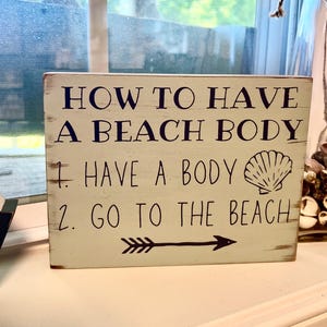 May include: A rectangular, off-white wooden sign with the text "HOW TO HAVE A BEACH BODY" in dark blue. Below, it reads "1. HAVE A BODY" and "2. GO TO THE BEACH" with a seashell graphic and an arrow.