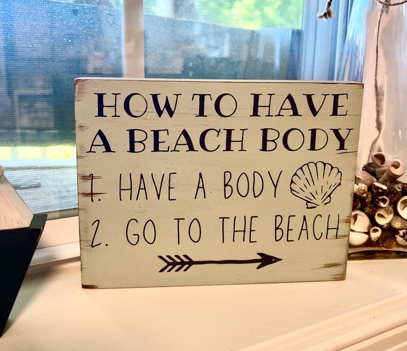 May include: A rectangular, off-white wooden sign with the text "HOW TO HAVE A BEACH BODY" in dark blue. Below, it reads "1. HAVE A BODY" and "2. GO TO THE BEACH" with a seashell graphic and an arrow.