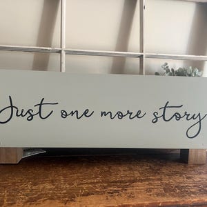 May include: A rectangular, light green wooden sign with the words "Just one more story" in black script. The sign is elevated on small, square wooden feet and sits on a wooden surface.
