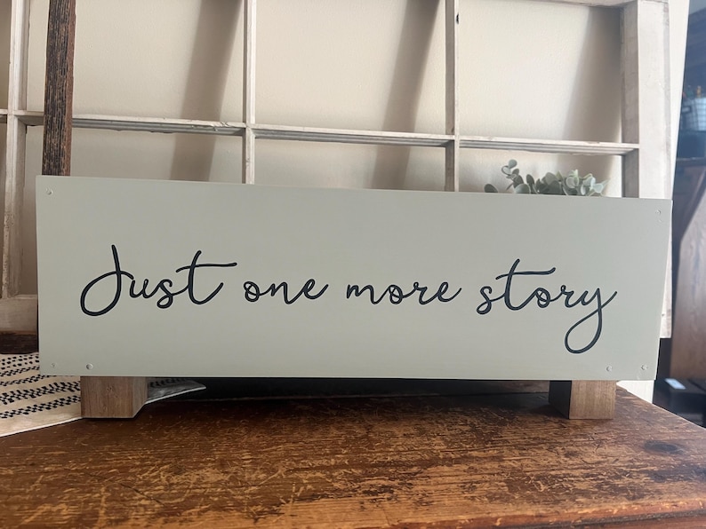 May include: A rectangular, light green wooden sign with the words "Just one more story" in black script. The sign is elevated on small, square wooden feet and sits on a wooden surface.
