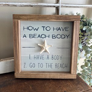 May include: Wooden framed sign with the text "HOW TO HAVE A BEACH BODY" and the steps "1. HAVE A BODY 2. GO TO THE BEACH". A white starfish is affixed to the sign. The frame is brown.