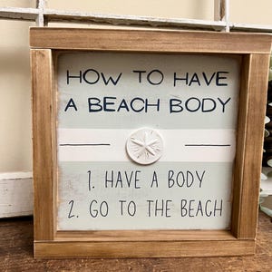 May include: Wooden framed sign with the text "HOW TO HAVE A BEACH BODY" and the steps "1. HAVE A BODY 2. GO TO THE BEACH". The sign has a light blue and white striped background and a white starfish accent.