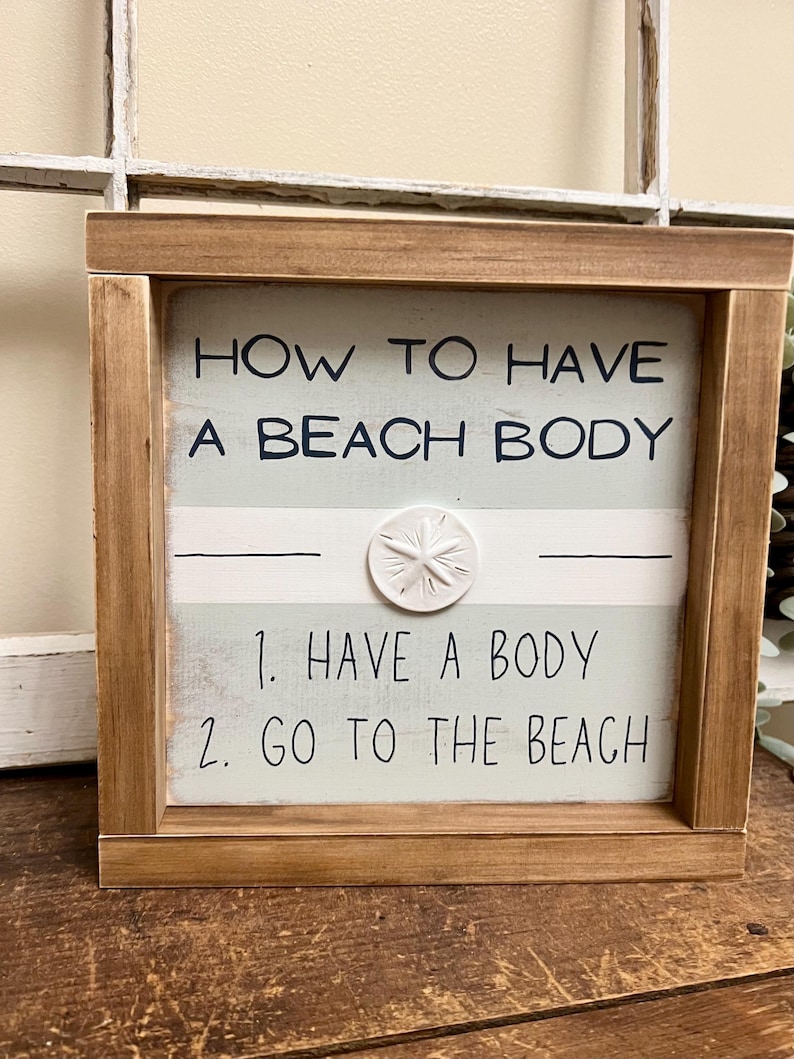 May include: Wooden framed sign with the text "HOW TO HAVE A BEACH BODY" and the steps "1. HAVE A BODY 2. GO TO THE BEACH". The sign has a light blue and white striped background and a white starfish accent.