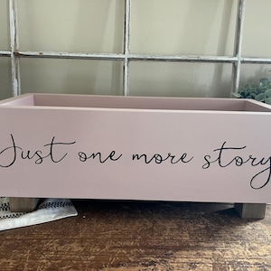 May include: A rectangular, light pink wooden box with the words "Just one more story" in black script. The box sits on four small, square wooden feet. The background includes a window and greenery.