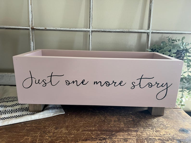 May include: A rectangular, light pink wooden box with the words "Just one more story" in black script. The box sits on four small, square wooden feet. The background includes a window and greenery.