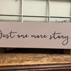 May include: A rectangular, light pink wooden box with the phrase "Just one more story" in black script. The box has dark brown wooden feet and rope handles. The background includes a window.